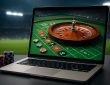 A laptop screen displaying a roulette wheel and chips, set against a blurred background of a stadium.